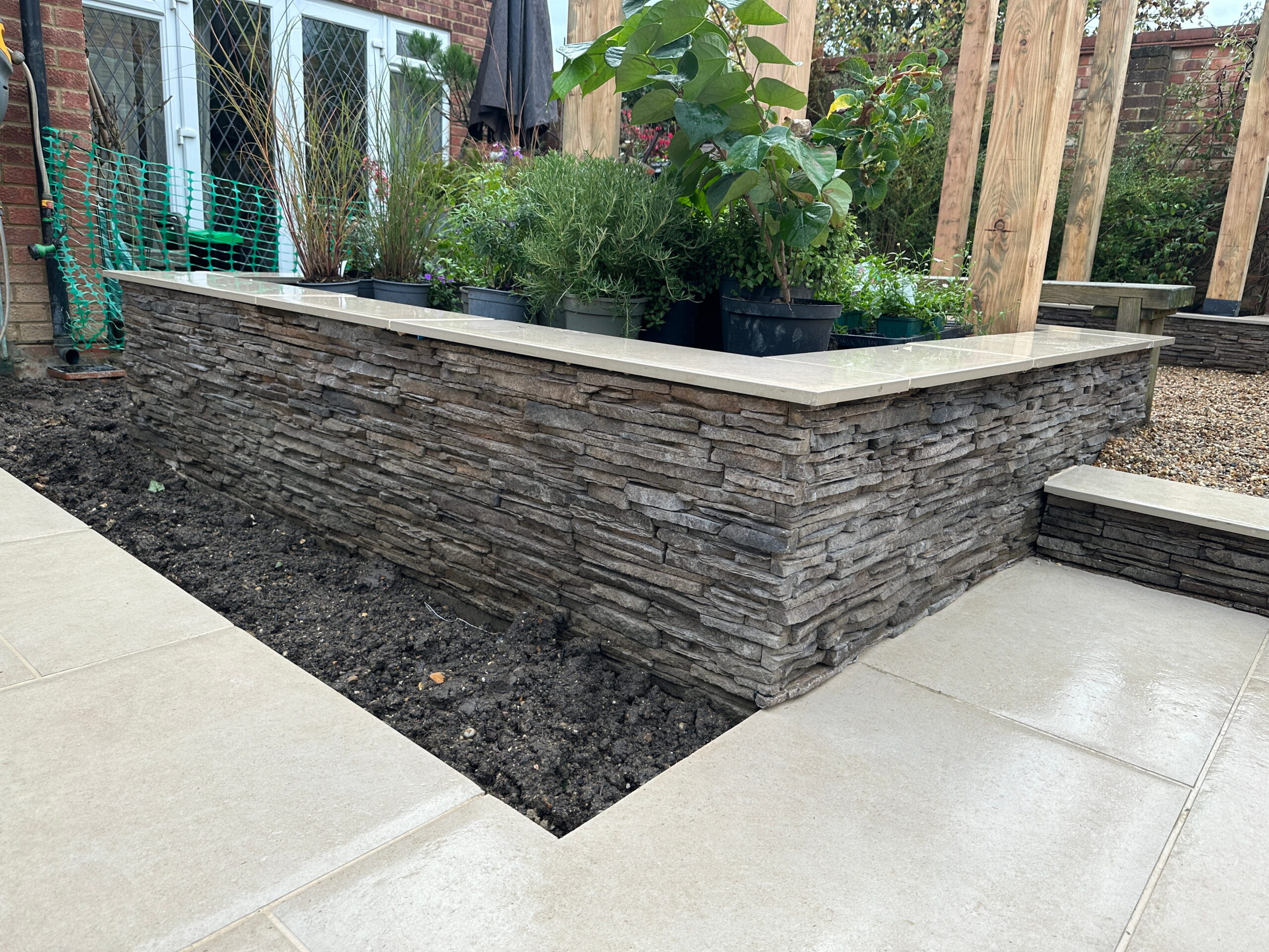 The image showcases a landscaped area with a stone wall planter, surrounded by various plants and newly laid paving