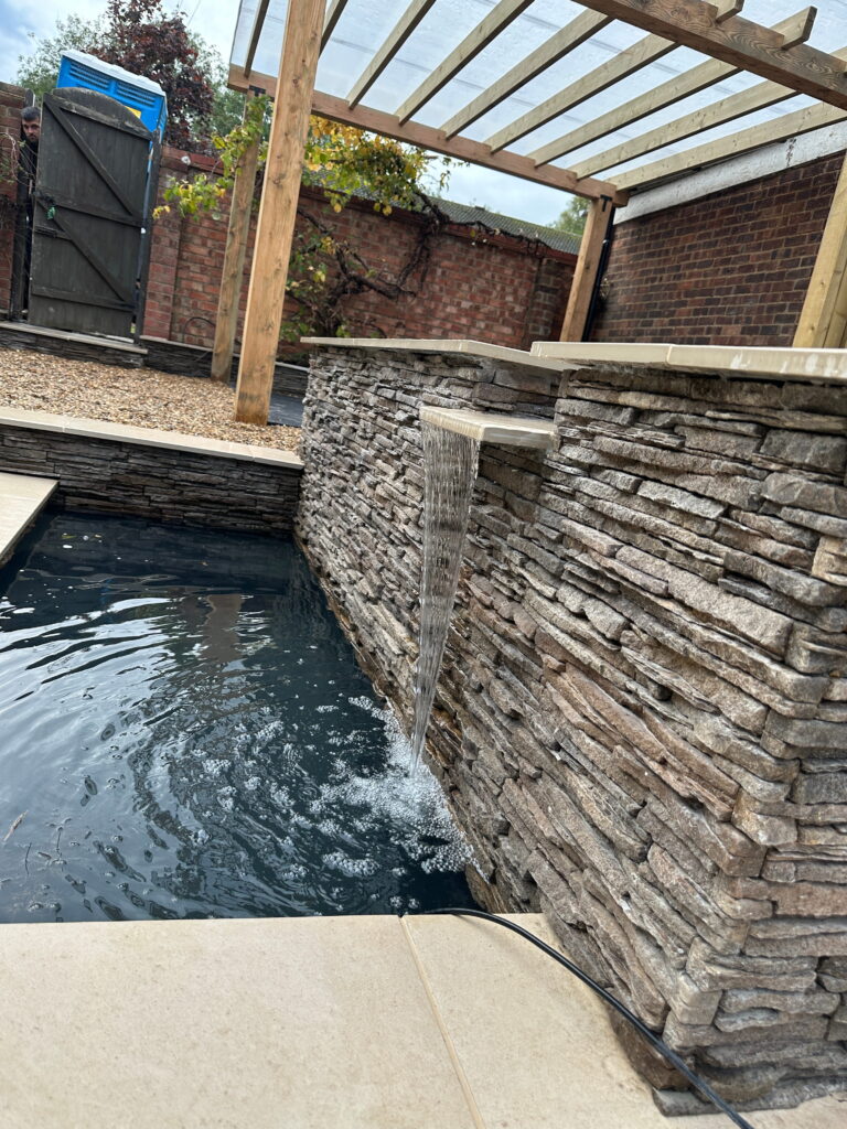 A sleek stone wall features a gentle water flow into a tranquil pool, with a pergola overhead and greenery nearby