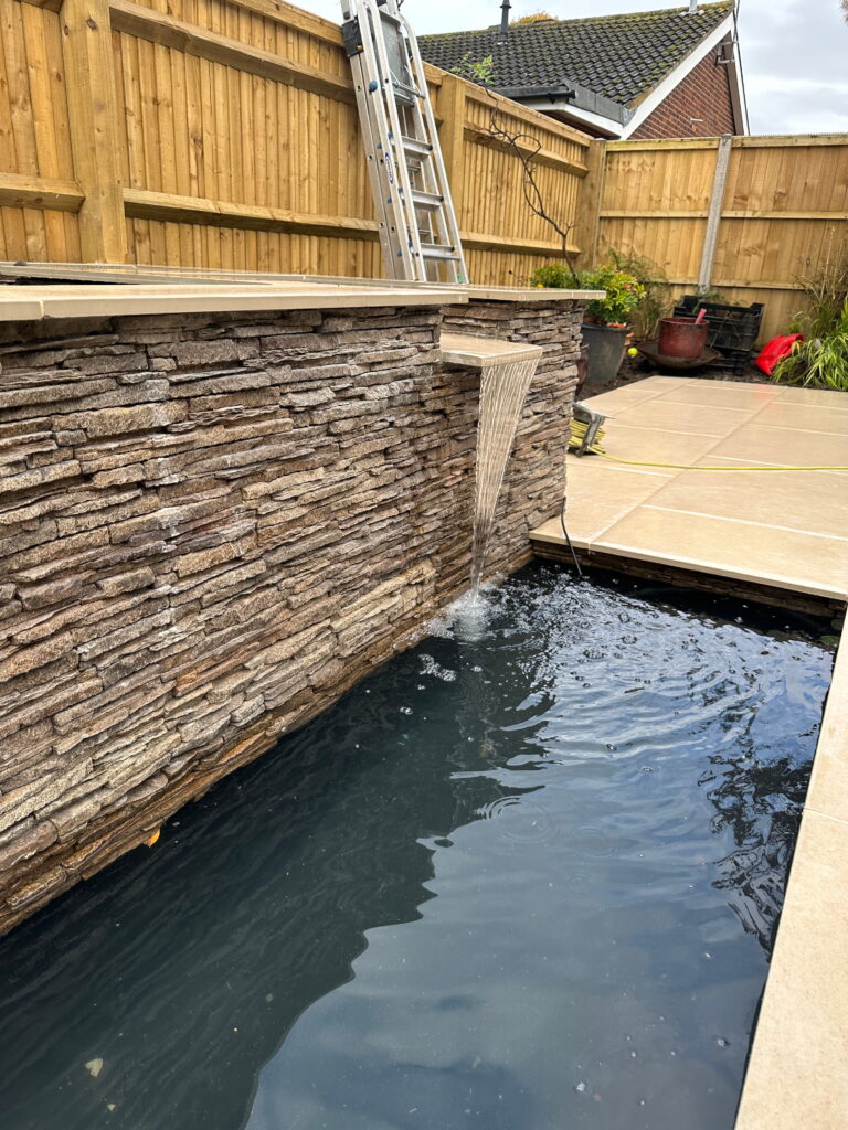 The image depicts a tranquil backyard setting featuring a stone wall with a water feature, cascading into a dark pool. A ladder leans against the wooden fence nearby