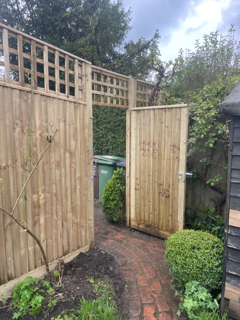 Wooden garden gate leading to a brick path, bordered by greenery and garden waste bins, under a cloudy sky