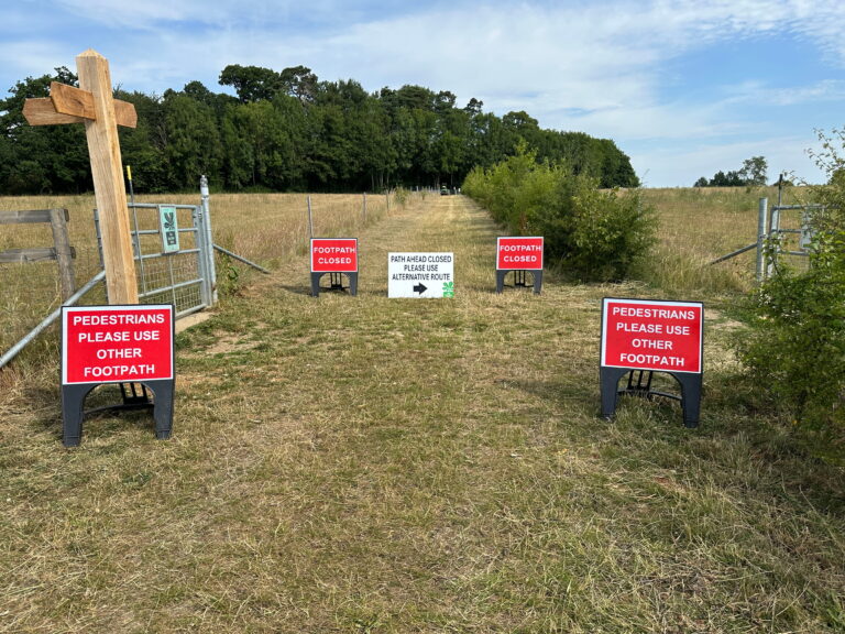 A closed footpath with multiple signs instructing pedestrians to use an alternate route, featuring a wooden cross in the foreground