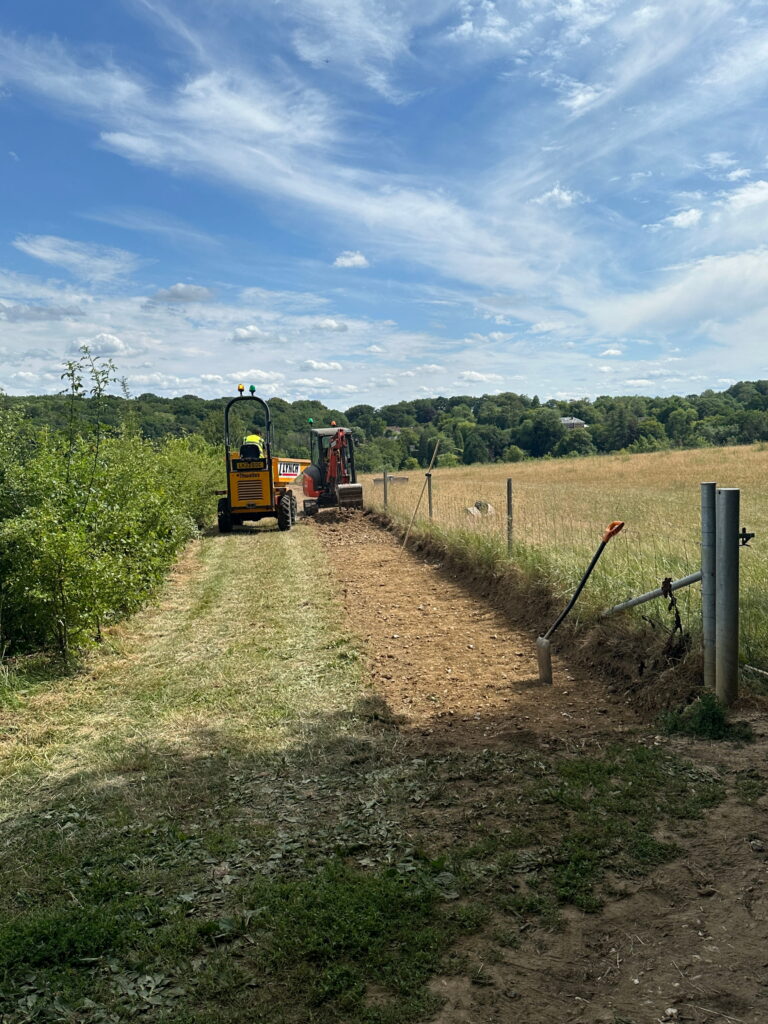 A sunny landscape features construction machinery working on a dirt path beside a grassy field, surrounded by trees
