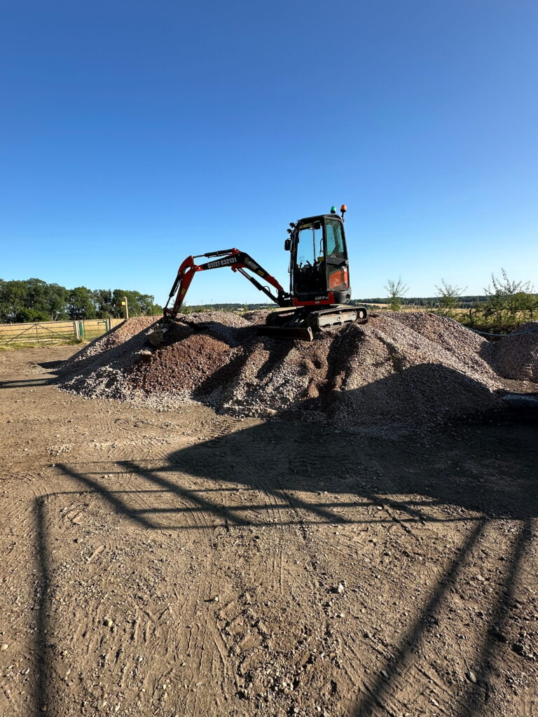 A compact excavator sits atop a gravel mound under a clear blue sky, surrounded by an open field