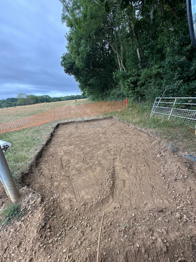 A cleared area of dirt, surrounded by orange fencing, near a field with trees in the background
