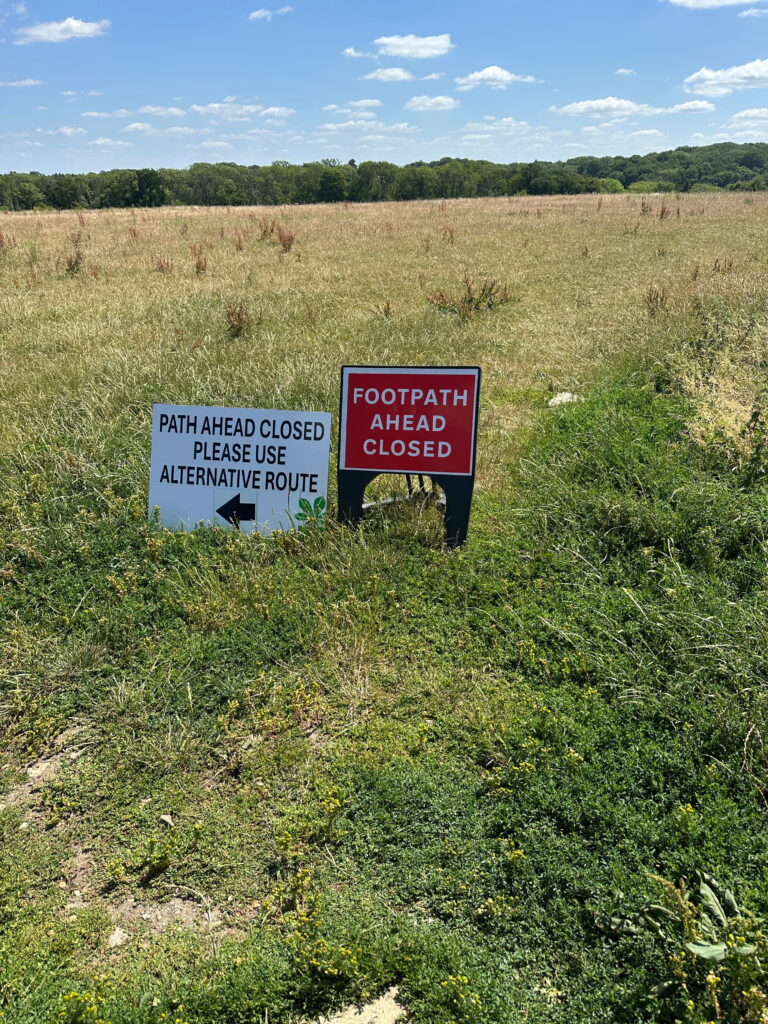 Two signs indicate path closures in a grassy field on a sunny day, suggesting an alternative route for pedestrians