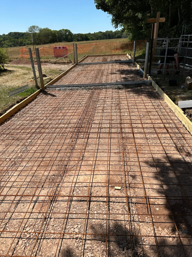 The image shows a construction site with a freshly laid foundation, featuring exposed rebar and gravel. A wooden framework outlines the area, with a clear blue sky in the background