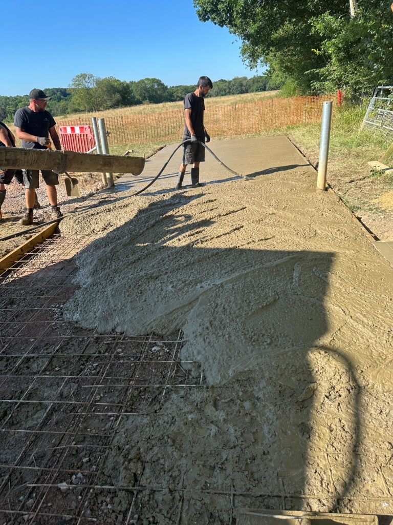Workers are pouring fresh concrete on a sunny day, smoothing it over a prepared area with visible rebar beneath