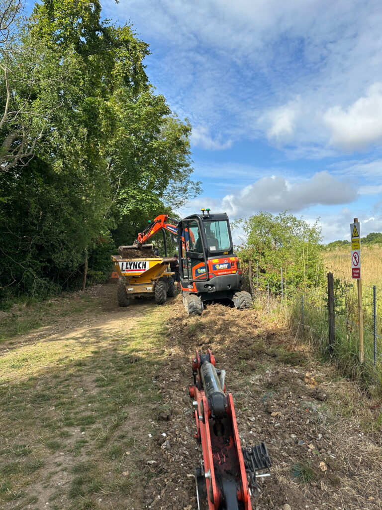 A construction site featuring a mini excavator and a dumper truck on a dirt path surrounded by trees and grass