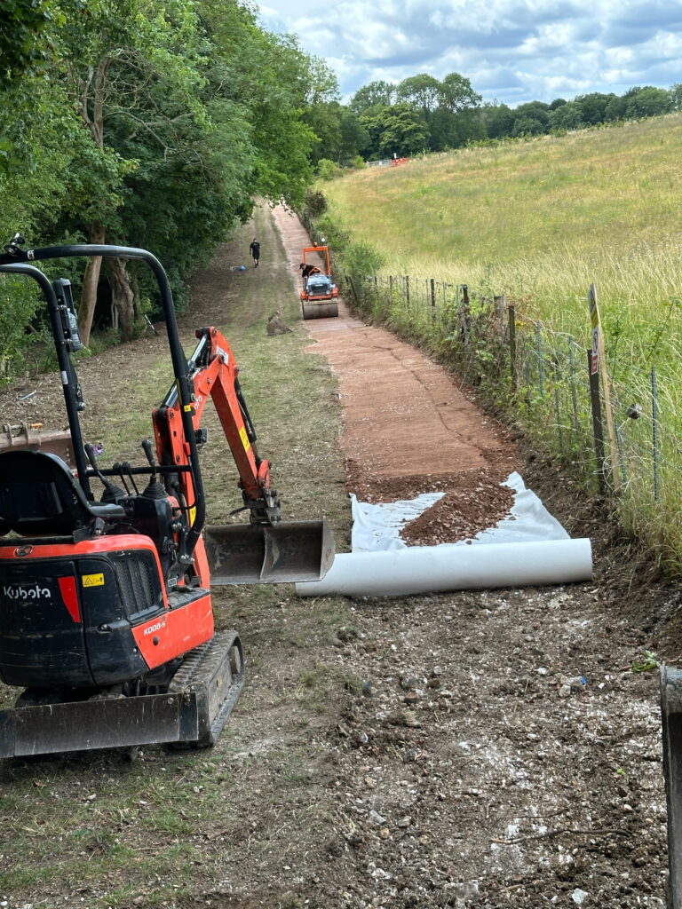 Construction equipment is actively working on a path, with a mini-excavator and roller applying materials on the ground