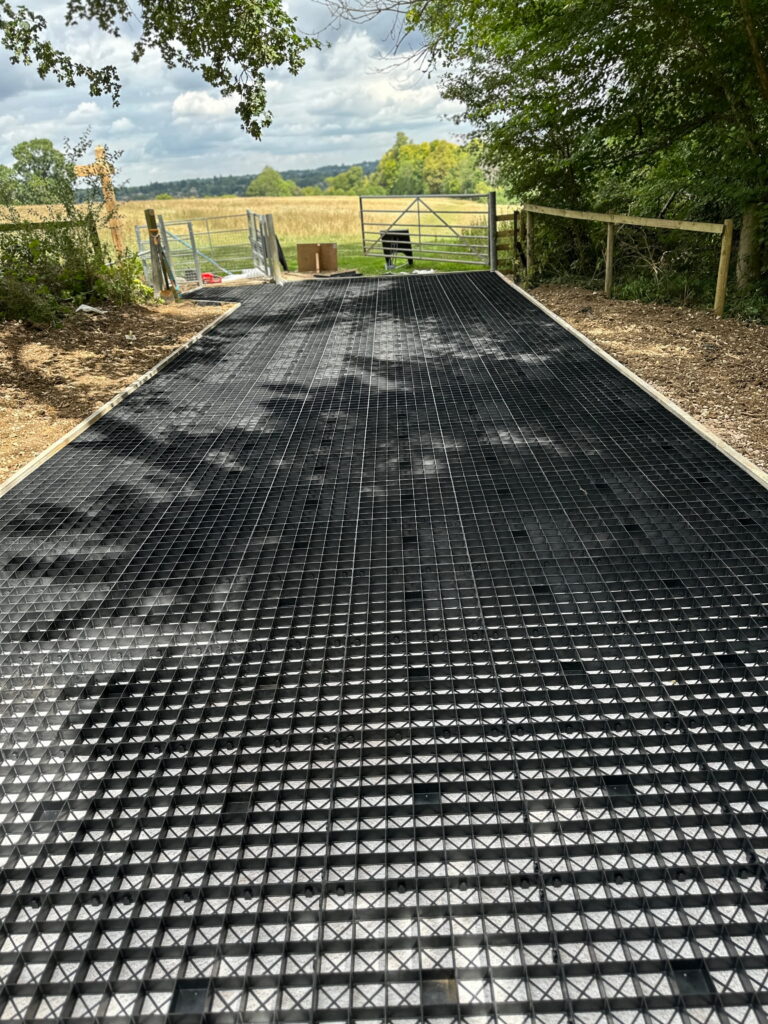 A textured black pathway leads toward a gate, surrounded by trees and fields under a partly cloudy sky