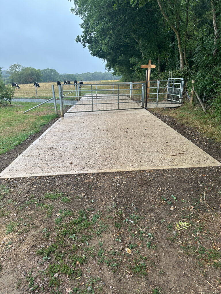 A gravel path leads to a gated entrance in a rural area, with cows grazing in the background under a cloudy sky