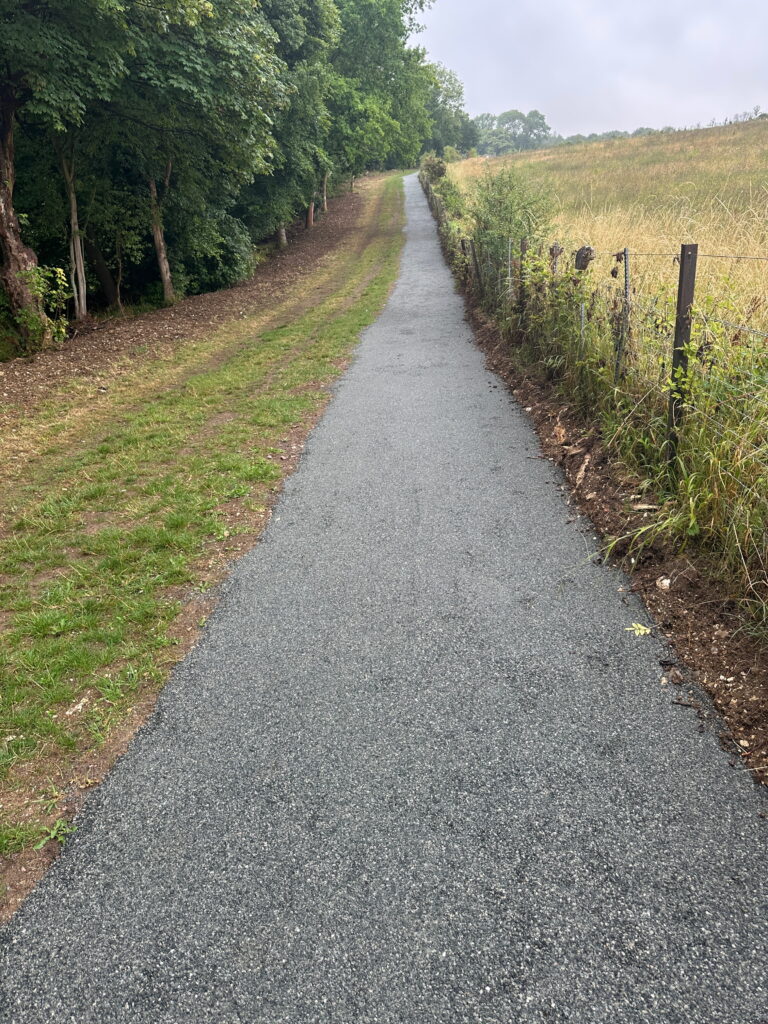 A paved path leads into a lush green area, bordered by trees on one side and fields on the other
