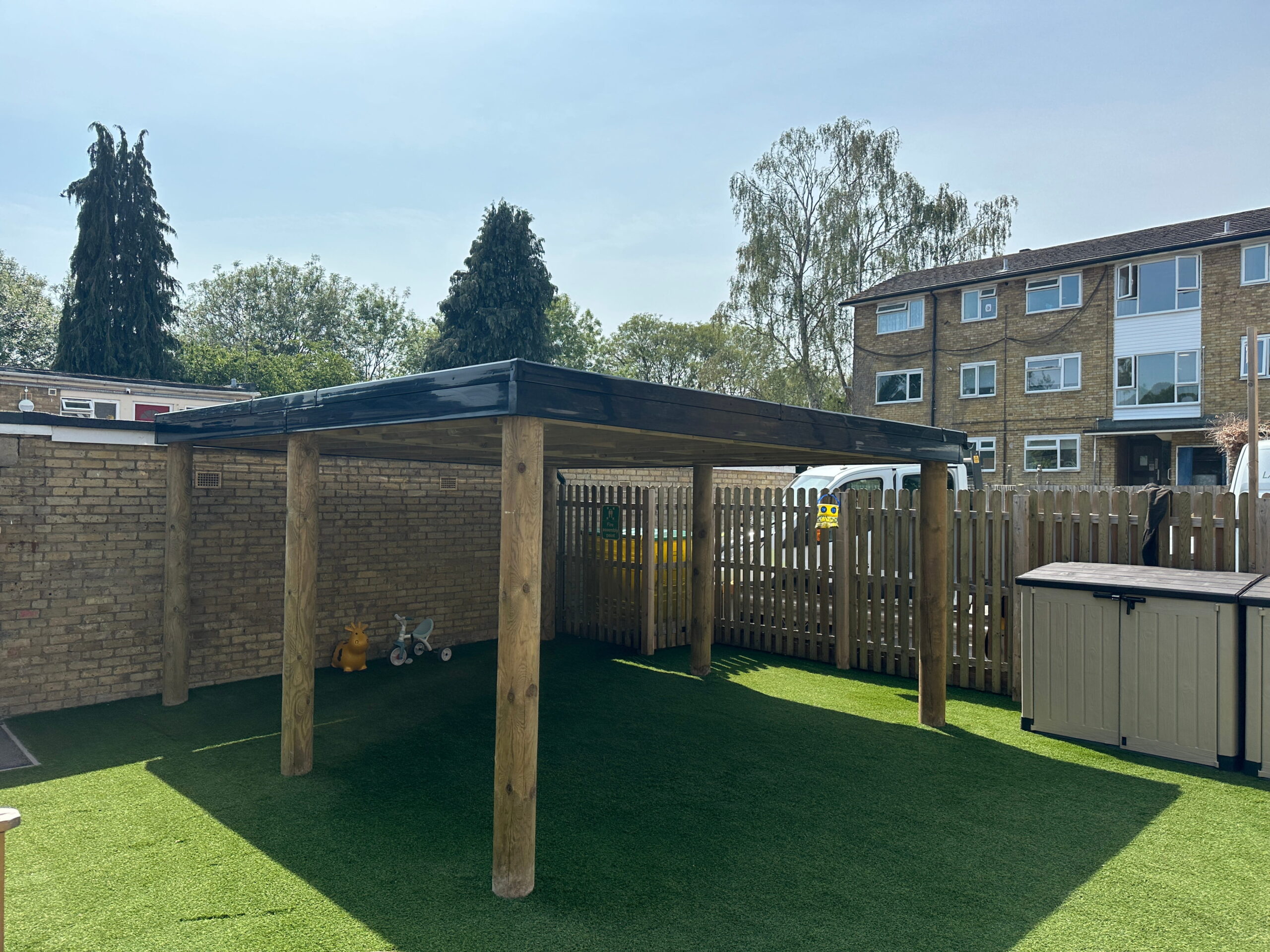 Wooden pergola in a sunlit backyard with artificial grass, surrounded by trees and a wooden fence. Perfect for outdoor relaxation