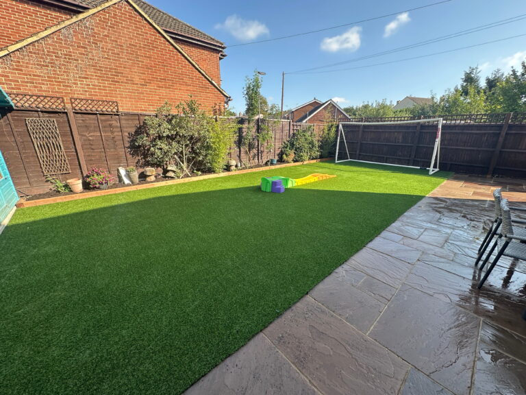 Bright backyard with artificial grass, play equipment, a soccer goal, and patio seating under a clear blue sky