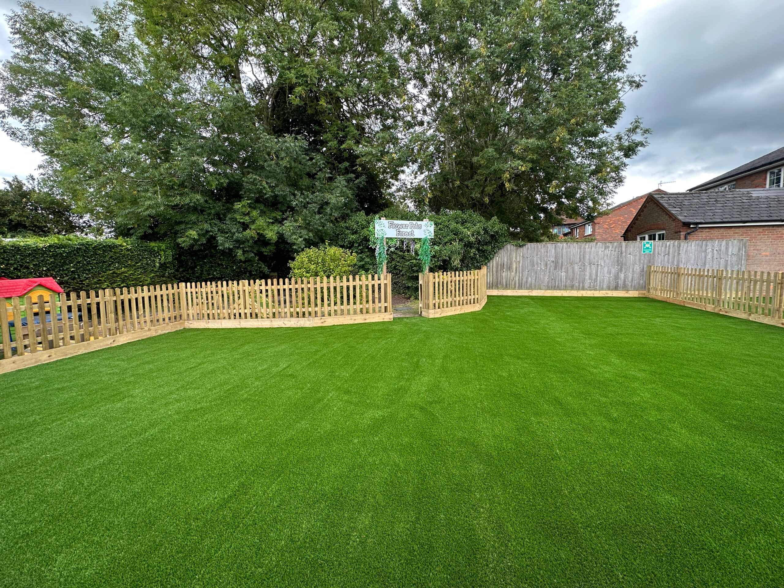 Lush green artificial grass playground with wooden fencing, surrounded by trees and a small colorful playhouse