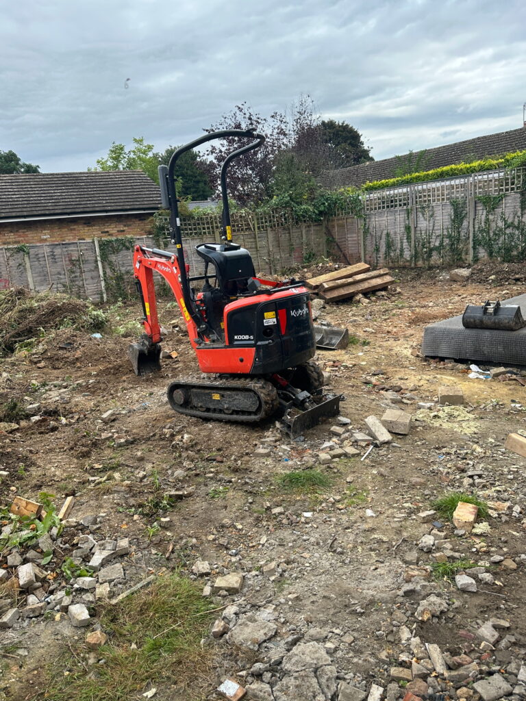 A Kubota mini excavator sits on a rubble-strewn site, surrounded by debris and overgrown vegetation, under a cloudy sky