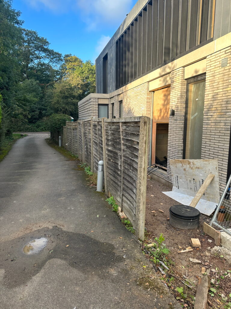A modern building beside a gravel path, featuring fencing, greenery, and construction materials scattered nearby
