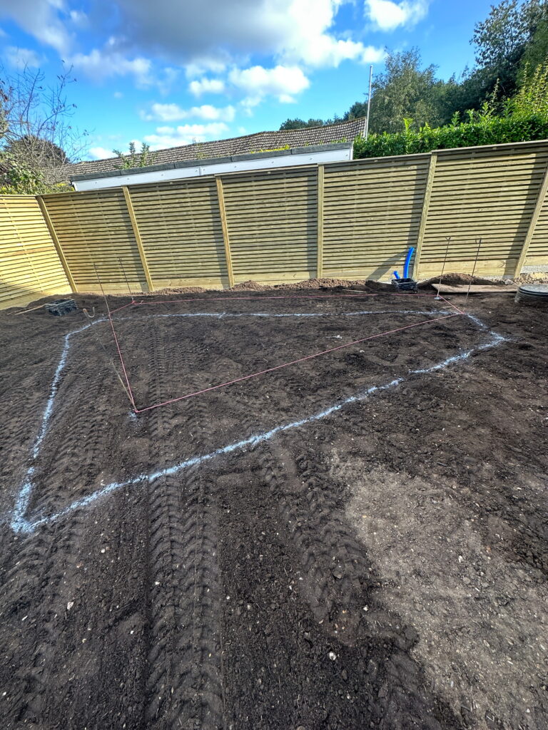 An outdoor area shows marked boundaries for construction, with dirt and tire tracks, surrounded by a wooden fence under a cloudy sky