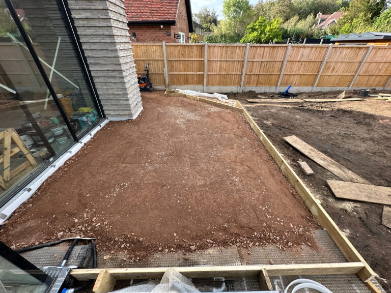 A construction site featuring red soil, a wooden border, and a modern building's glass wall. Tools and materials are nearby