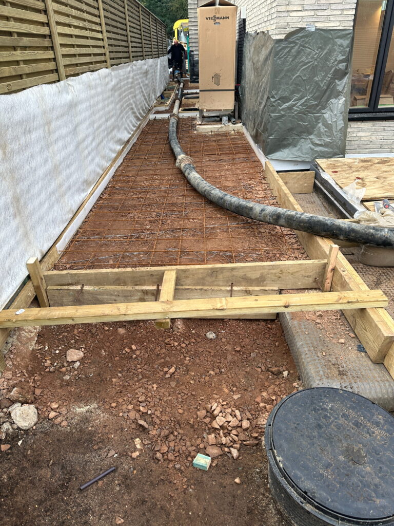 The image shows a construction site with a concrete foundation in progress. There's a wooden frame, rebar mesh, and a hose for concrete pouring