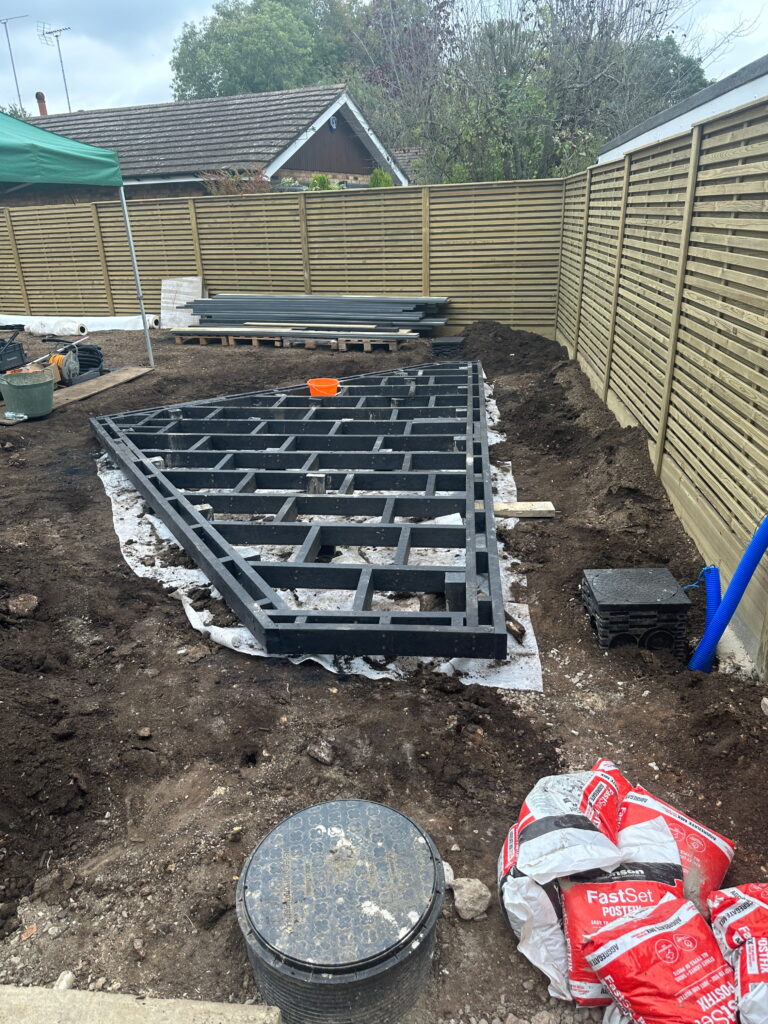 The image shows a construction site with a black frame on soil, tools, materials, and a green tent in the background