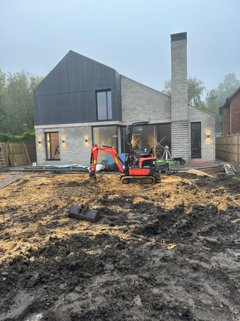 A modern house under construction, featuring a mini excavator on a muddy site with scattered construction materials