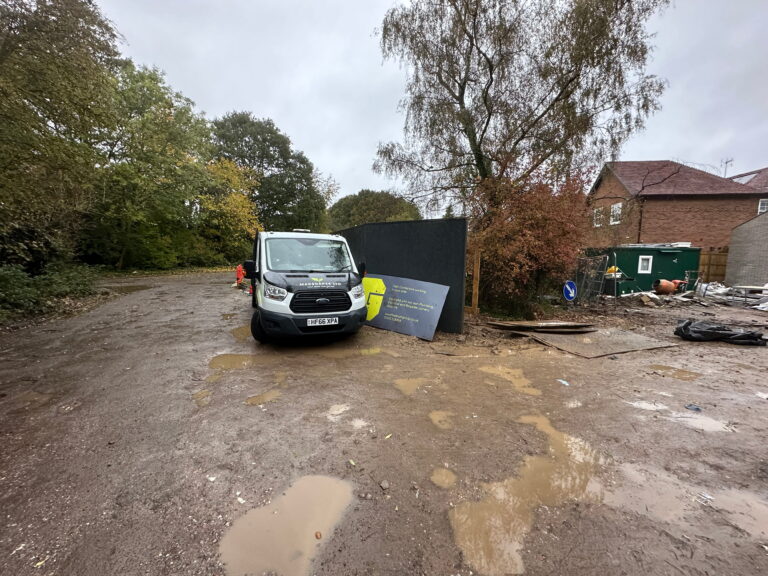 A muddy construction site with a parked white van, a black wall, and warning signs amidst trees and debris