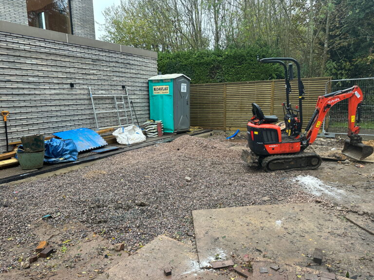 A construction site features gravel, a mini excavator, scaffolding, and a portable restroom, surrounded by a wooden fence