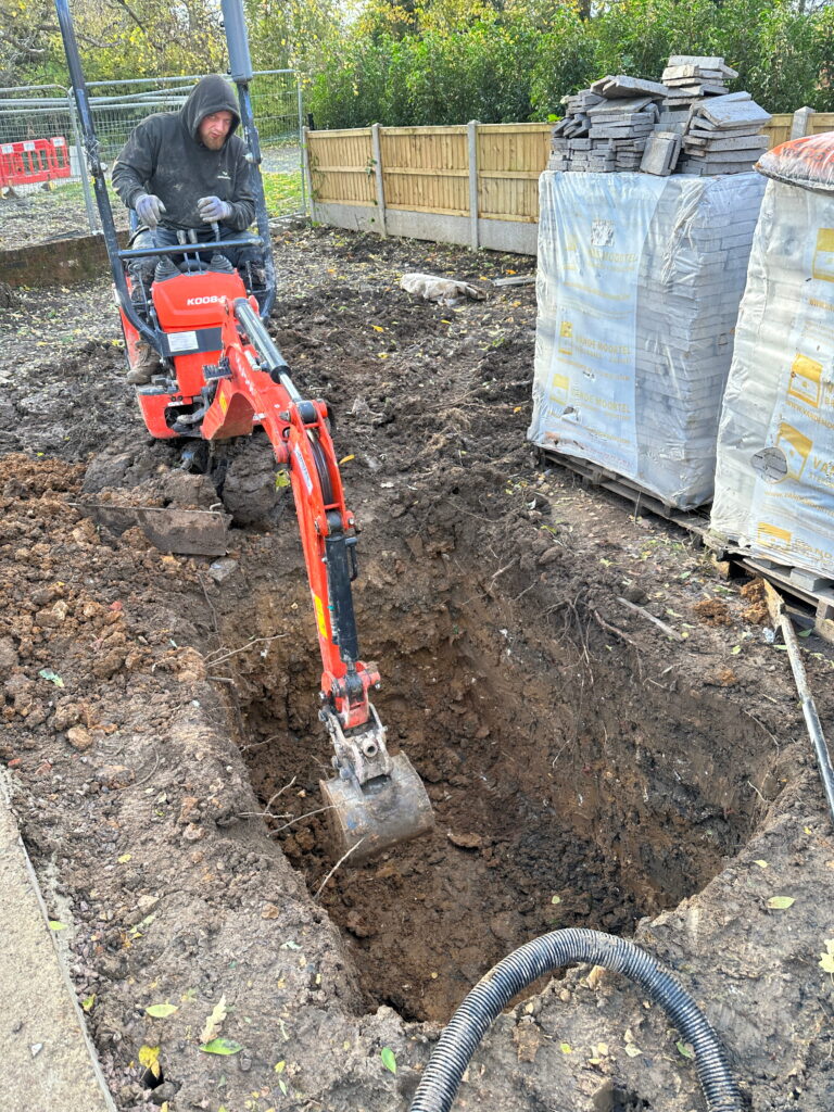 A worker operates a small excavator, digging a deep hole in a construction site, surrounded by soil and materials