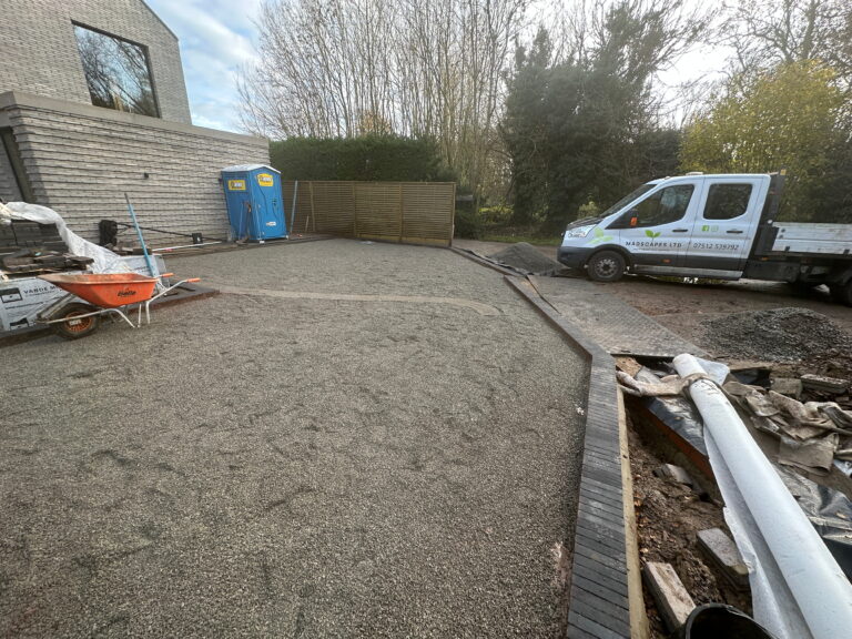 A partially completed landscaping site features gravel, a wheelbarrow, a work truck, and portable toilets among greenery