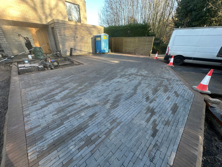 A newly laid brick driveway features varying shades, with construction tools and a portable restroom in the background. Safety cones are placed around the area