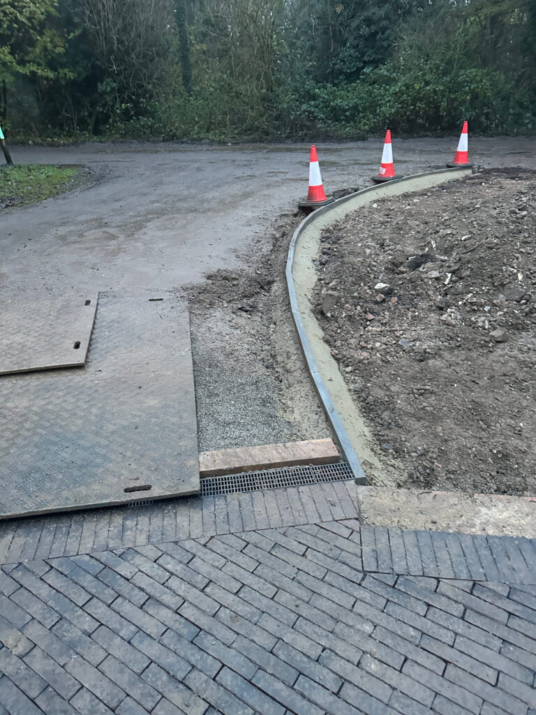 A construction area with a curved path, gravel, mats, and traffic cones, surrounded by greenery
