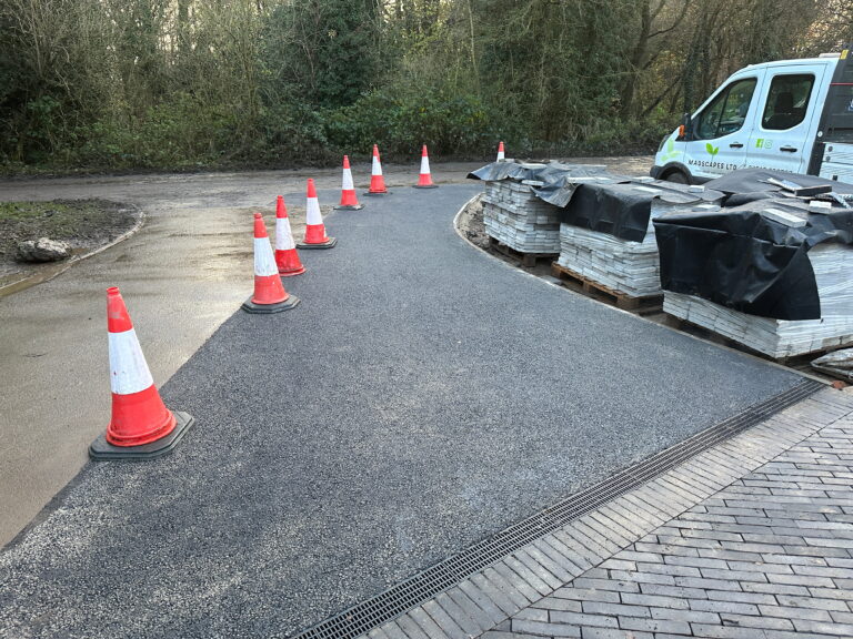 A newly paved pathway is lined with orange traffic cones, with pallets of covered materials nearby, surrounded by greenery