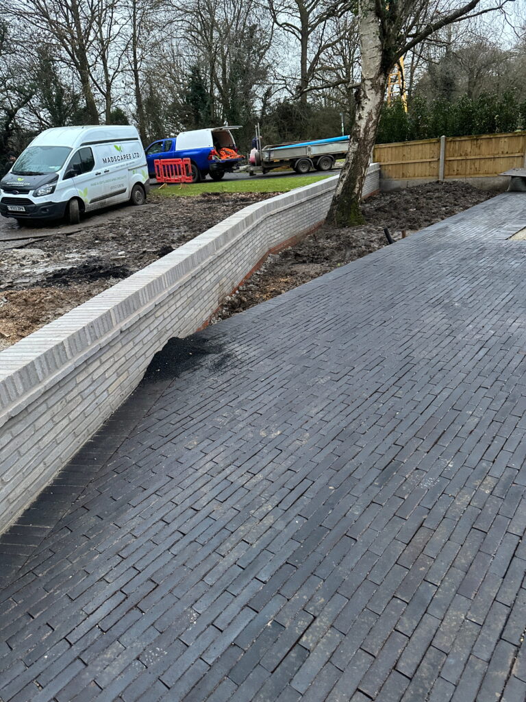 The image shows a construction site with a newly paved brick pathway, vehicles, and landscaping materials in a wooded setting