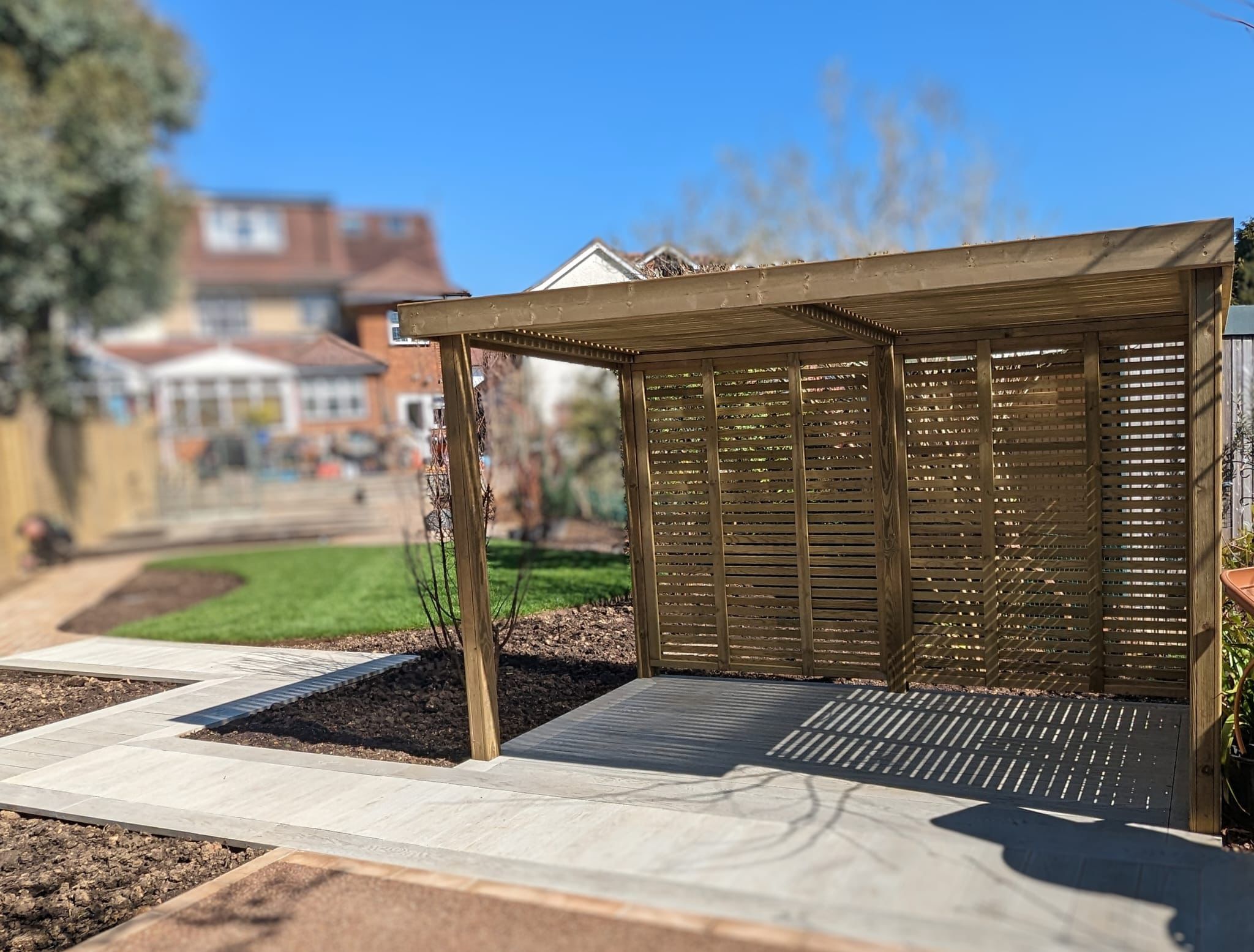A wooden garden shelter with slatted walls, set against a sunny backdrop of green grass and a blue sky