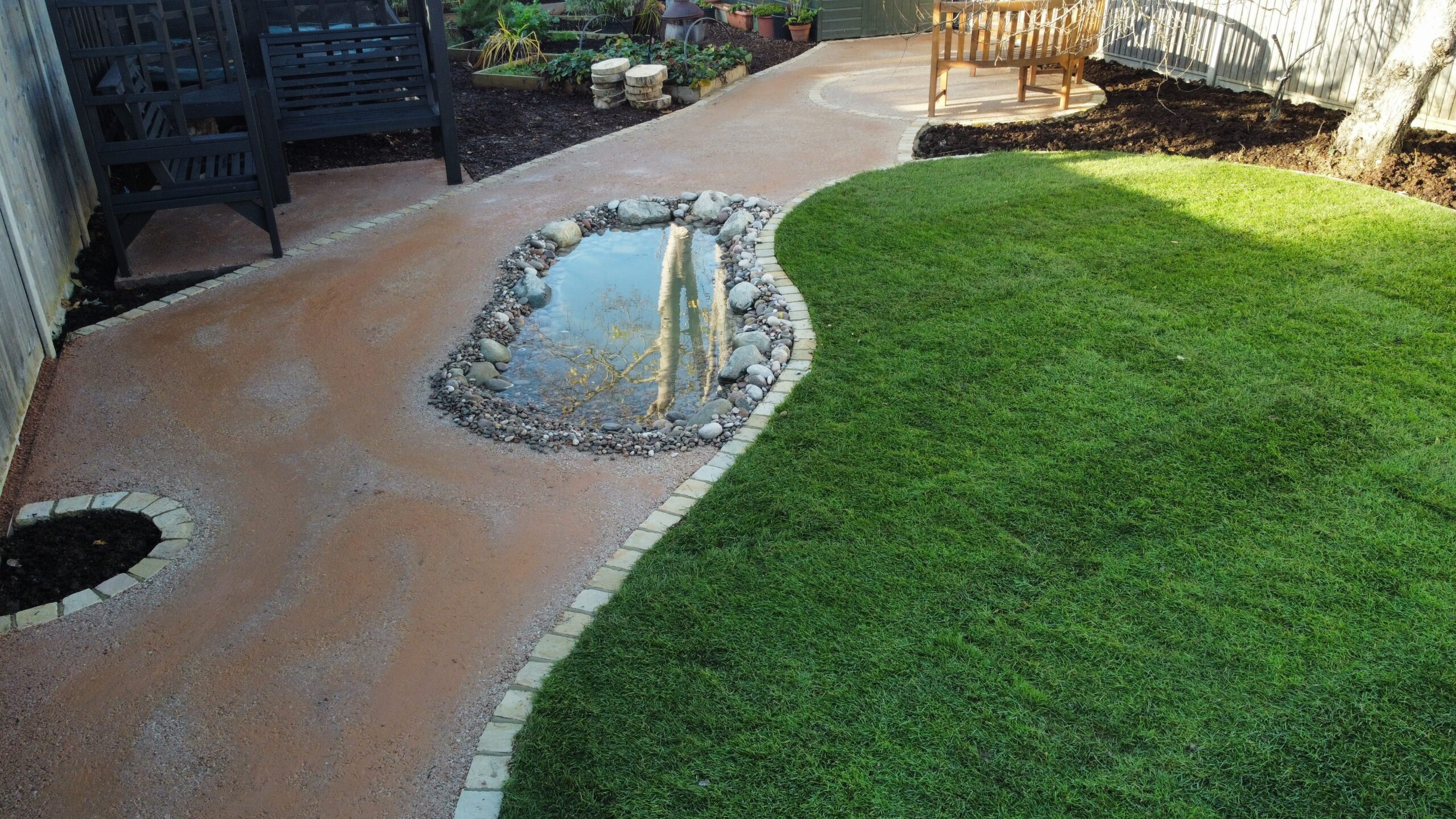 Inviting backyard with curvy pathway, serene pond, lush green grass, and cozy seating for relaxation and gardening