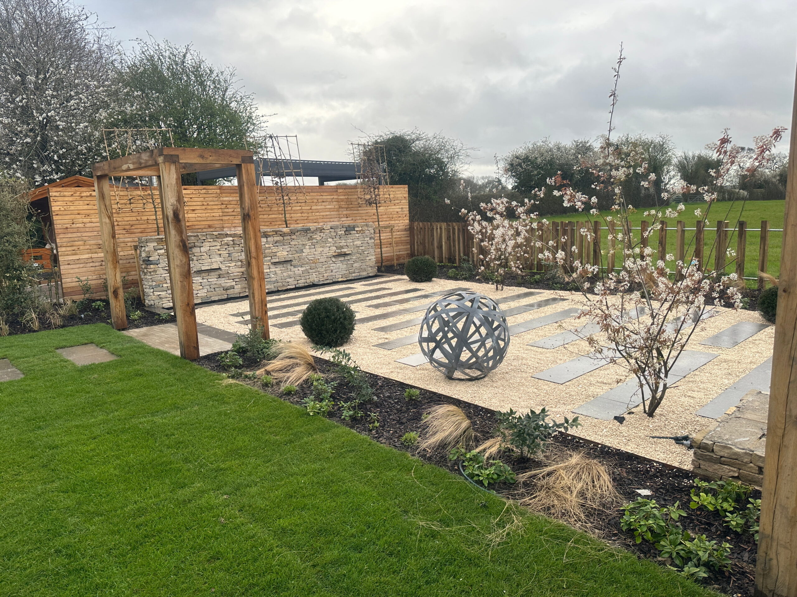 Modern garden design featuring a stone patio, ornamental globe sculpture, blooming trees, and landscaped greenery. Perfect for outdoor relaxation