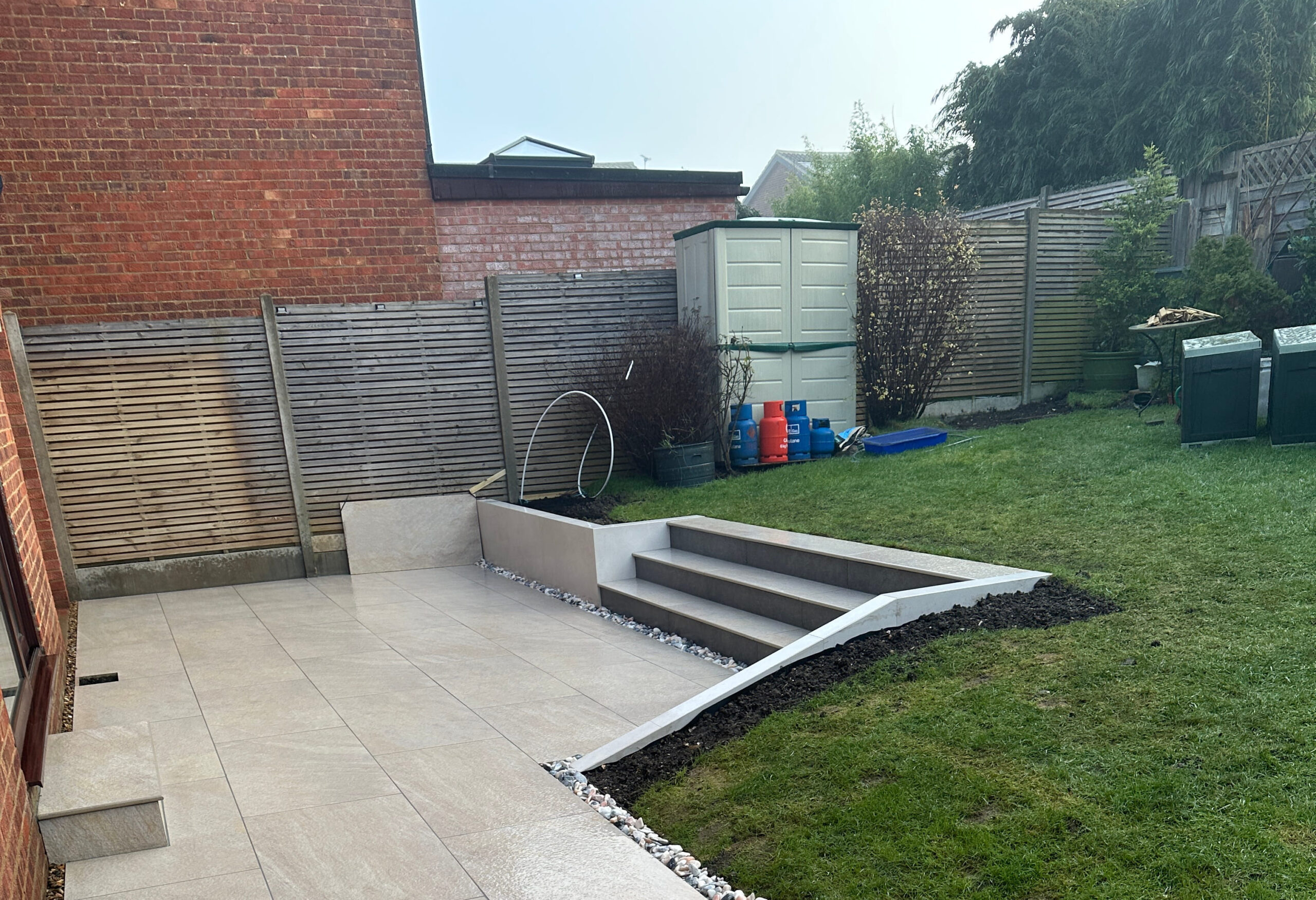 The image shows a backyard with modern paving, steps, a storage shed, and a small lawn area