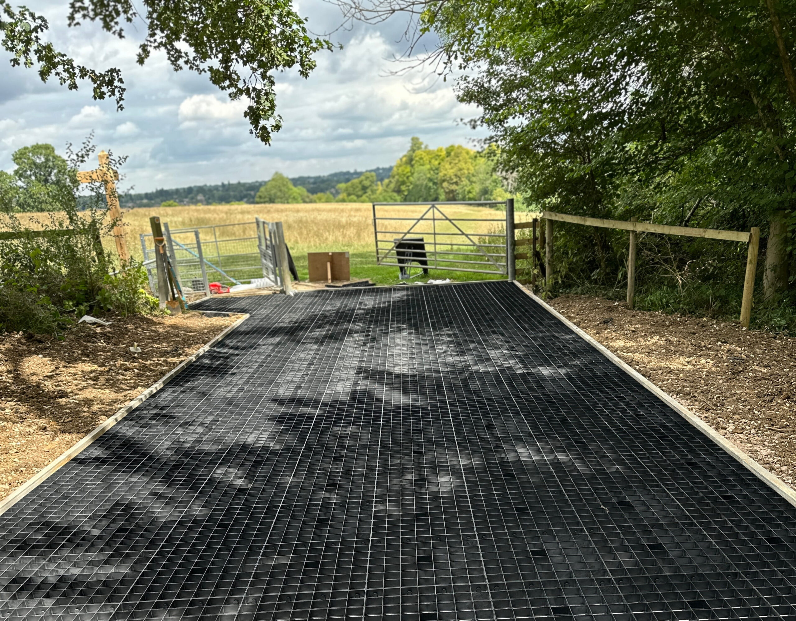 The image shows a grid-patterned black pathway leading to a gate, surrounded by trees and fields