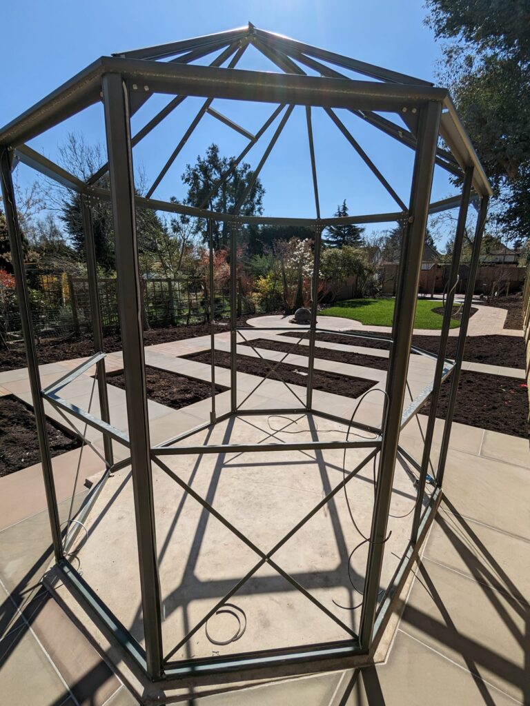 Metal gazebo frame under a clear blue sky, surrounded by landscaped garden and freshly laid pavers