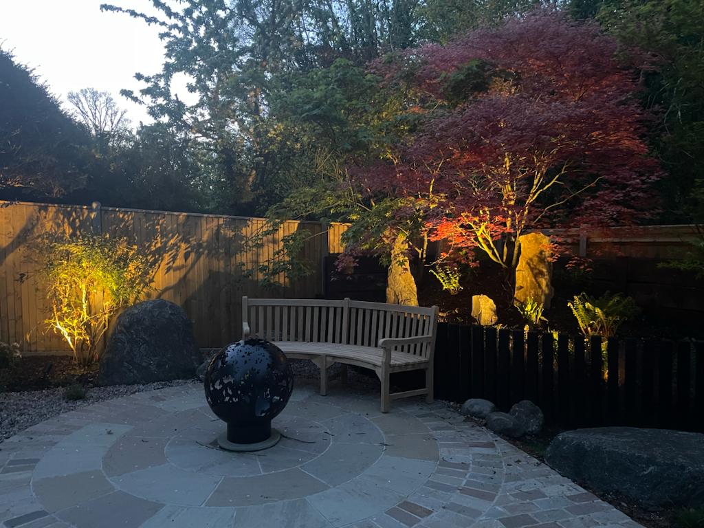 Serene garden at dusk featuring a maple tree, stone seating, and a decorative sphere, illuminated by soft lighting
